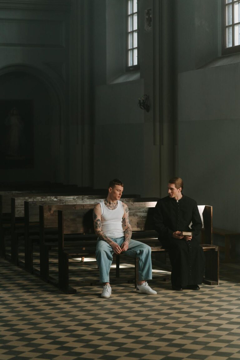A tattooed man sitting with a priest in a dimly lit church, symbolizing confession and repentance.