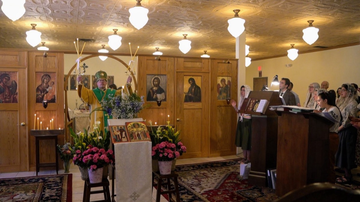 A bishop in green and gold vestments raises candlesticks in blessing before the icon screen, while a choir of women in veils sings at a stand on the right