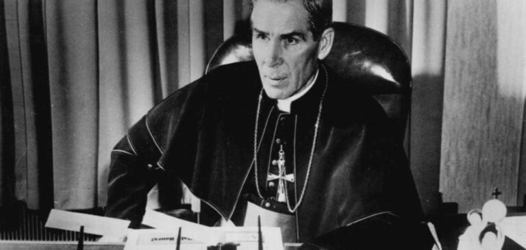 Bishop Fulton J. Sheen at his desk in 1956, in cassock and shoulder cape with pectoral cross, papers before him.